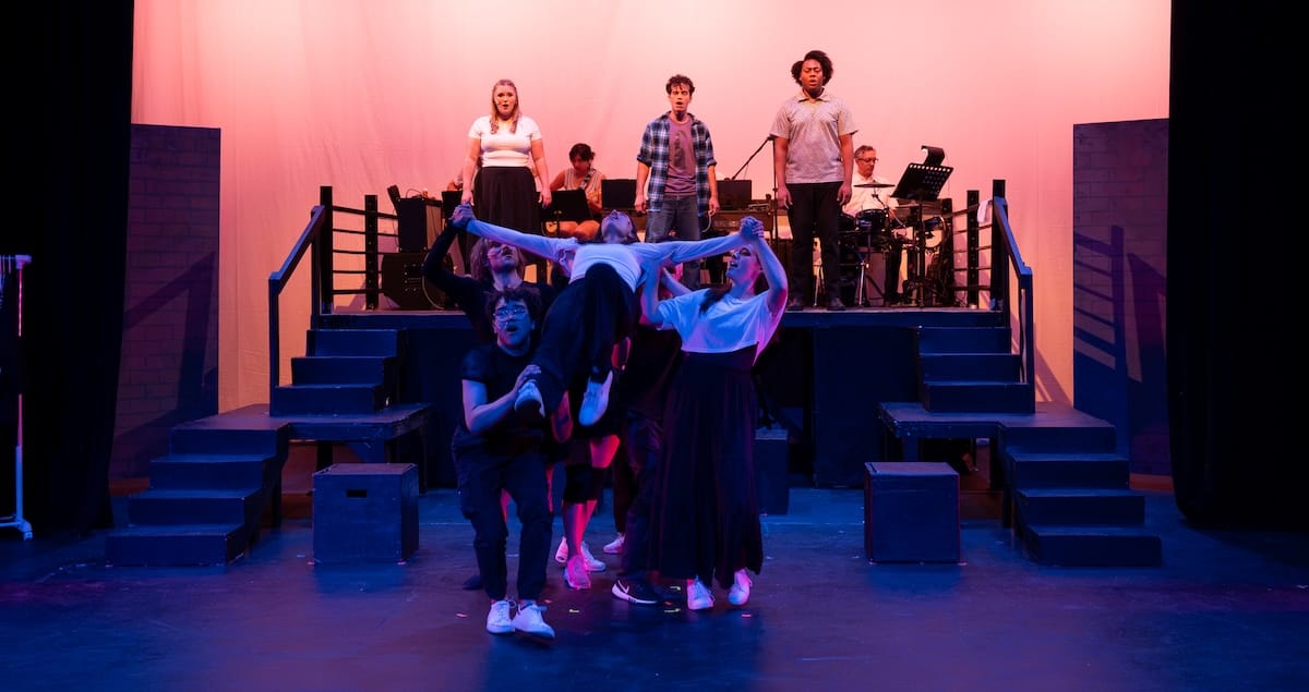 A group of people lift another person as three people sing on a platform behind them