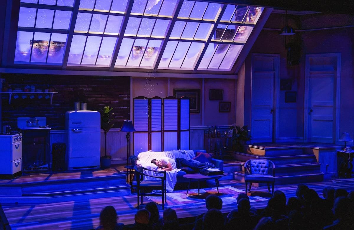 A man sleeps on the couch as snow falls on him through a hole in the ceiling in a scene from "Barefoot in the Park"