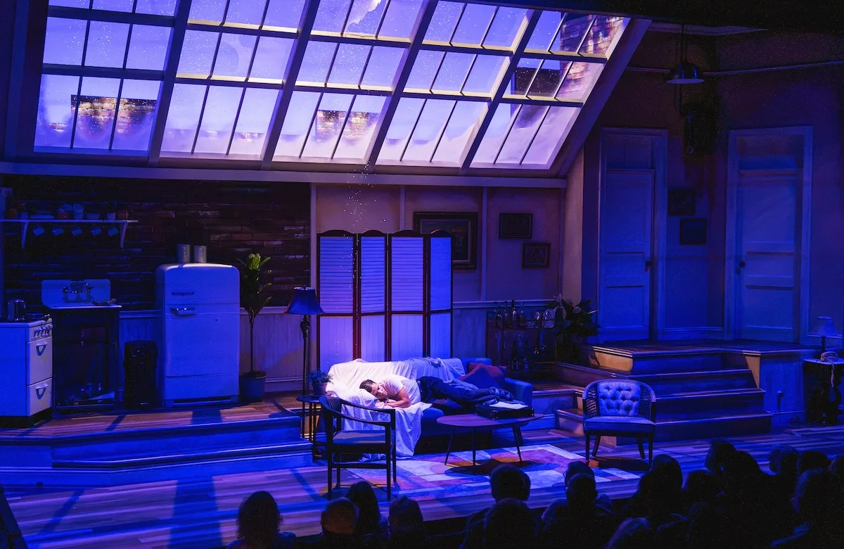 A man sleeps on the couch as snow falls on him through a hole in the ceiling in a scene from "Barefoot in the Park"